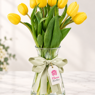 Tulipanes amarillos con follaje en florero de vidrio, moño verde y etiqueta Flores Bellas.