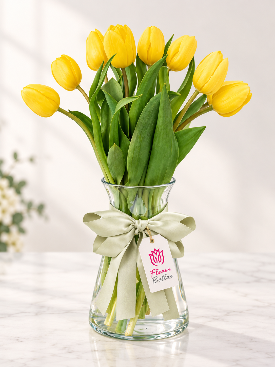 Tulipanes amarillos con follaje en florero de vidrio, moño verde y etiqueta Flores Bellas.